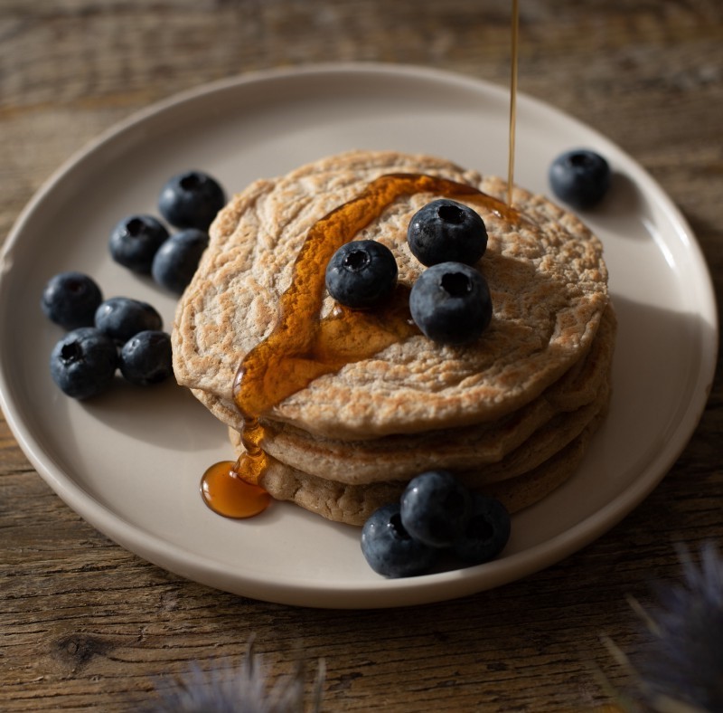 3 whole-meal pancakes with a drizzle of maple syrup and blueberries on a beige plate.