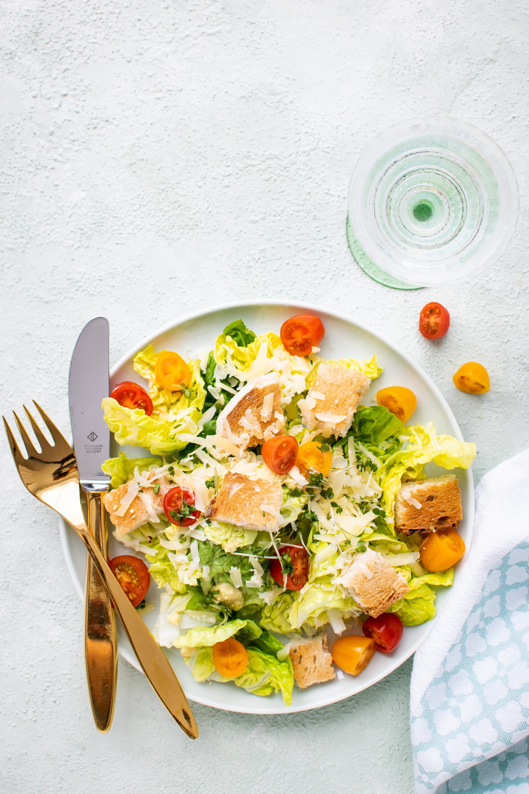 A colorful plate of salad consisting of lettuce, cherry tomatoes, croutons and vegan shredded cheese, served with a fork and knife.